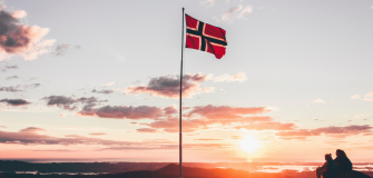 two people sat by a Norway flag on top of a mountain during sunset