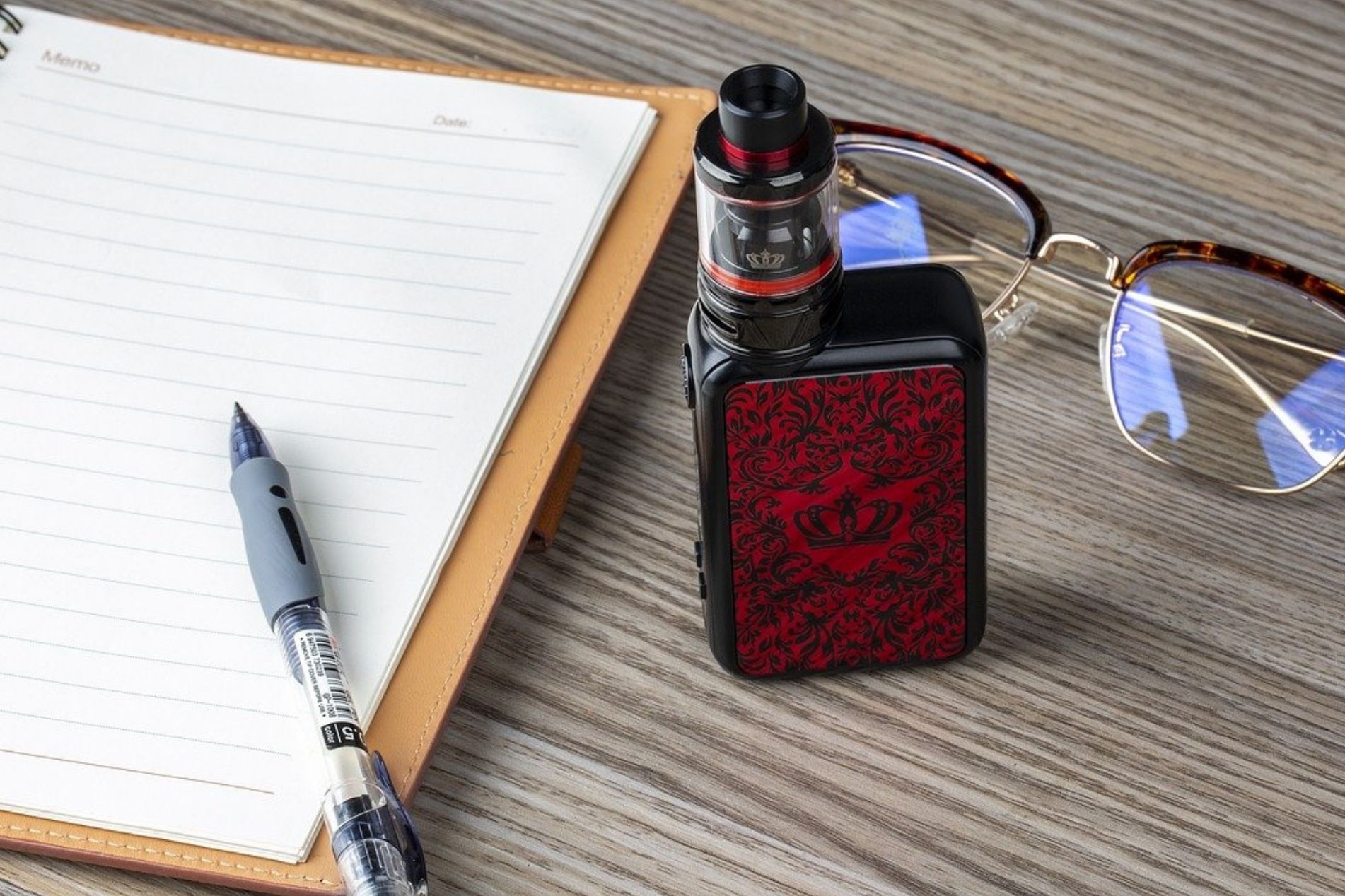 A red and black vape sits ona. wooden desk next to a notepad and a pair of glasses