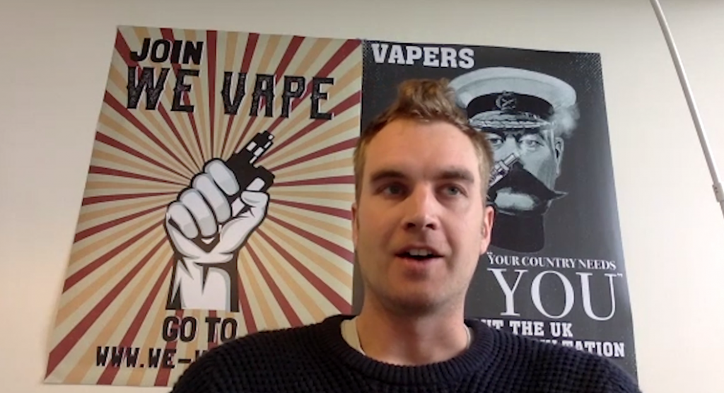 A man sits in front of a poster stating 'join we vape' featuring a hand holding a vape in it