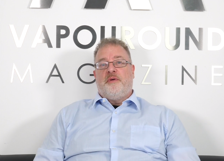 A man sits in a white room wearing a light blue shirt. A silver sign in the background reads Vapouround Magazine